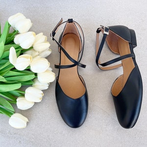 Könnte beinhalten: Ein Paar schwarze Ballerinas aus Leder mit gekreuzten Riemen. Die Ballerinas haben eine spitze Zehenpartie und einen niedrigen Absatz.