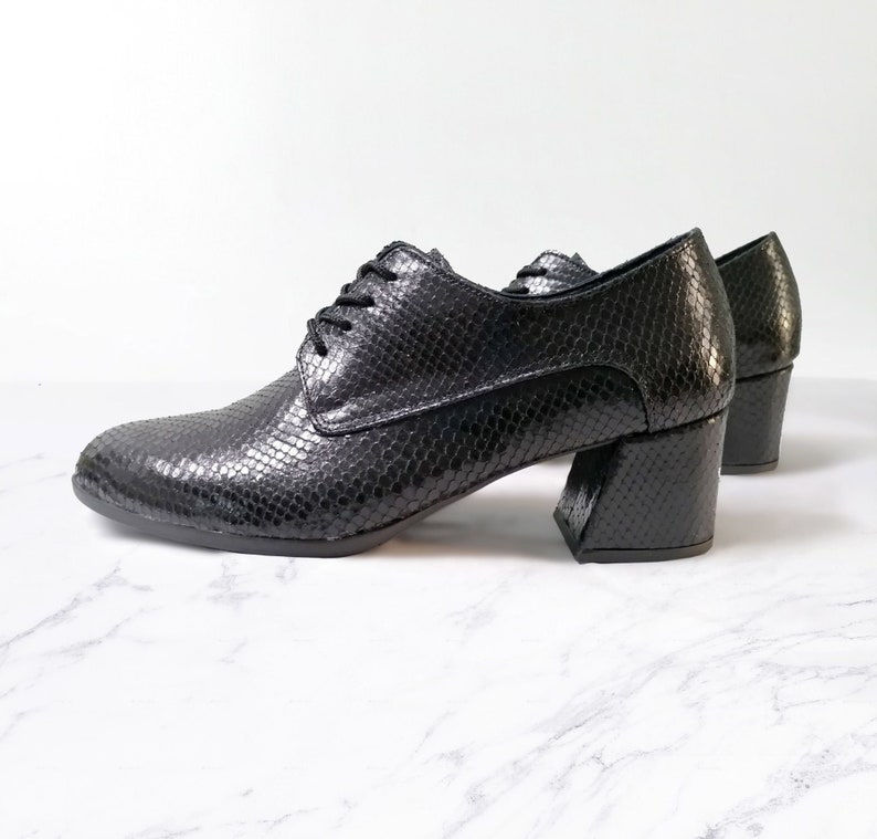 May include: Black leather oxford shoes with a snakeskin texture. The shoes have a lace-up design and a chunky, block heel. The heels are approximately 3 inches (7.6 cm) high. The shoes are set against a white background.
