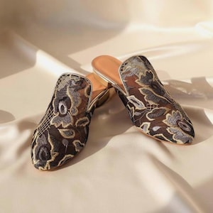Könnte beinhalten: Ein Paar elegante Mules mit offenem Rücken. Die Schuhe haben eine schwarze florale Spitze mit goldenen und silbernen Akzenten. Das Innere ist hellbraun und die Schuhe haben einen kleinen Absatz.