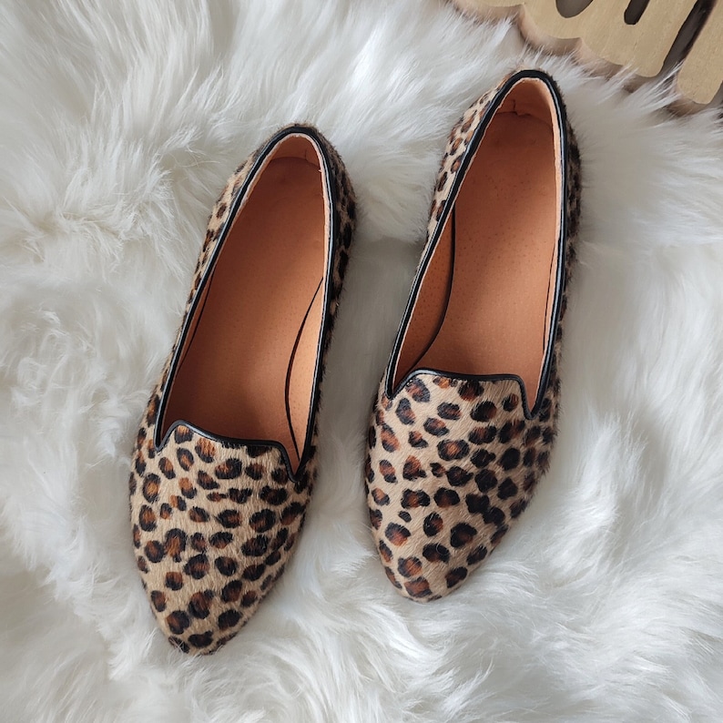 May include: A pair of leopard print slip-on shoes with black trim. The shoes are made of a soft, fuzzy material and have a pointed toe.