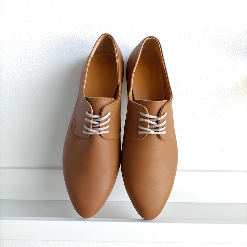 May include: A pair of brown leather oxford shoes with white laces. The shoes have a classic design with a pointed toe and a low heel. The shoes are displayed on a white surface.