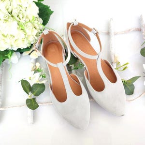 May include: A pair of light gray suede T-strap flats with ankle straps and buckle closures. The shoes have a pointed toe and a brown insole. The image is styled with white flowers and greenery, suggesting a bridal or special occasion theme.