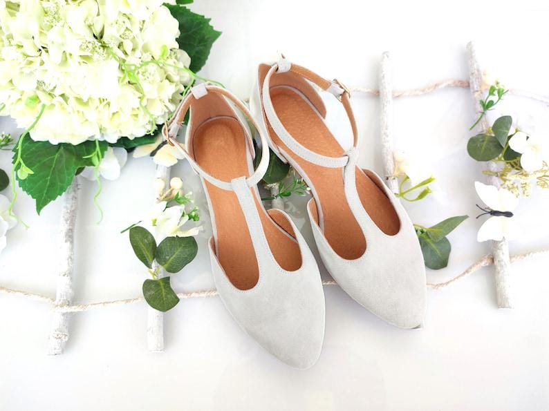 May include: A pair of light gray suede T-strap flats with ankle straps and buckle closures. The shoes have a pointed toe and a brown insole. The image is styled with white flowers and greenery, suggesting a bridal or special occasion theme.