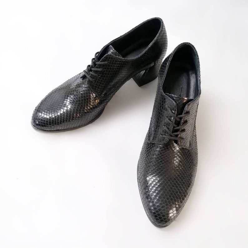 May include: Pair of black leather oxford shoes with a textured snakeskin pattern. The shoes have a lace-up closure and a block heel. The shoes are displayed on a white background.