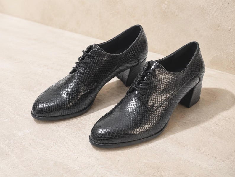 May include: A pair of black, textured leather oxfords with a snakeskin pattern. The shoes have a lace-up design and a block heel. The shoes are displayed on a neutral-colored surface.