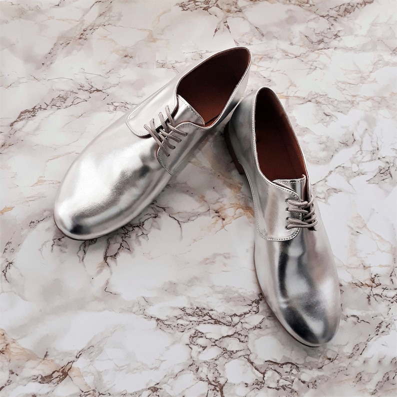 May include: A pair of silver leather oxfords with lace-up closures. The shoes are on a white marble background.