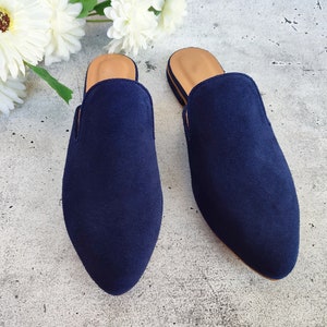 May include: Two navy blue suede slip-on mules with a pointed toe and a tan insole. The shoes are displayed on a gray surface with white flowers in the background. The mules are a stylish footwear option.