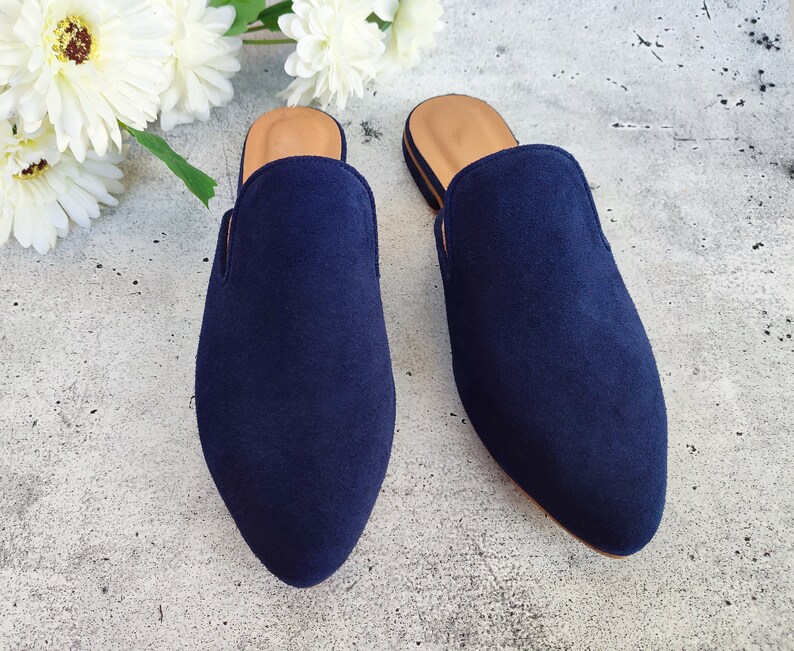 May include: Two navy blue suede slip-on mules with a pointed toe and a tan insole. The shoes are displayed on a gray surface with white flowers in the background. The mules are a stylish footwear option.