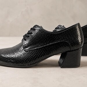 May include: Black, textured leather oxfords with a low, block heel. The shoes have a lace-up closure and a rounded toe. The leather has a snakeskin pattern. The shoes are set against a neutral background.