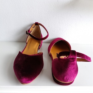 May include: A pair of burgundy velvet ankle strap flats. The shoes have a pointed toe and a low heel.