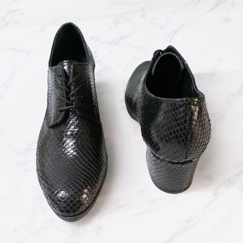 May include: Pair of black leather shoes with a snakeskin pattern. The shoes have a lace-up design and a small heel. The shoes are displayed on a white marble surface.