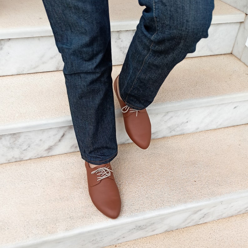 May include: Brown leather dress shoes with white laces. The shoes are worn with dark blue denim jeans. The image shows the shoes on a staircase with a marble finish.