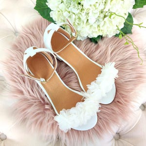 May include: A pair of white sandals with a flower embellishment on the straps. The sandals have a low heel and are made of a soft, comfortable material.