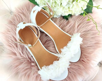 Puede incluir: Un par de sandalias blancas con un adorno floral en las tiras. Las sandalias tienen un tacón bajo y están hechas de un material suave y cómodo.