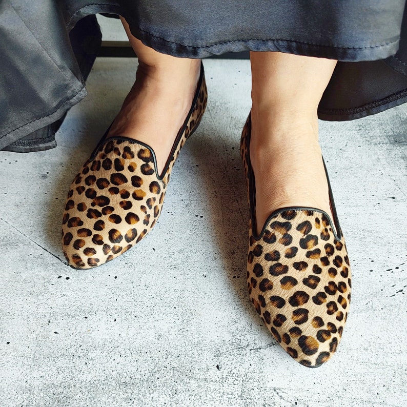 May include: A pair of leopard print flats with black trim. The shoes are made of a soft, fuzzy material and have a pointed toe.