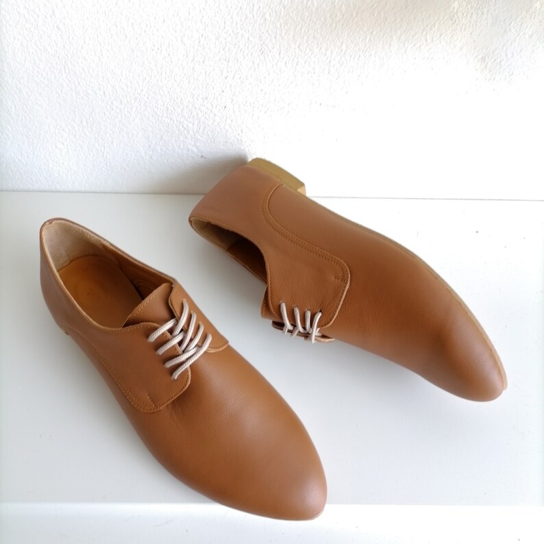 May include: A pair of brown leather shoes with white laces. The shoes have a classic design with a rounded toe and a low heel. The shoes are displayed on a white surface.