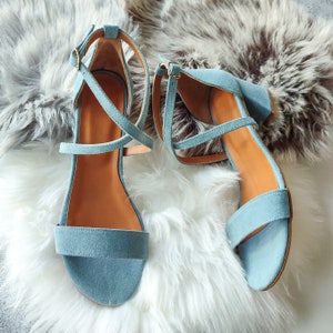 May include: A pair of light blue suede heeled sandals with ankle straps. The sandals have a criss-cross strap design and are shown on a white fluffy surface.