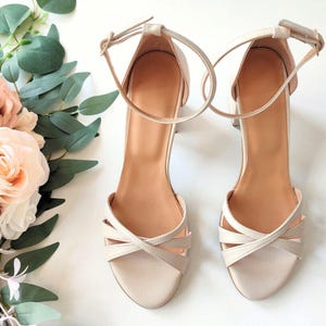 May include: A pair of cream-colored block heel sandals featuring an ankle strap with a buckle closure and a crisscross strap design across the open toe. The interior of the shoes is a tan color, and the exterior is a smooth, solid cream finish.
