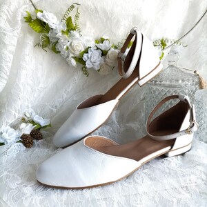 May include: A pair of white leather ankle strap flats with a pointed toe. The shoes are on a white lace fabric background.