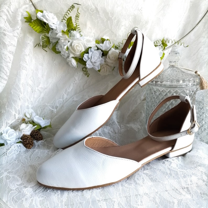 May include: A pair of white leather ankle strap flats with a pointed toe. The shoes are on a white lace fabric background.