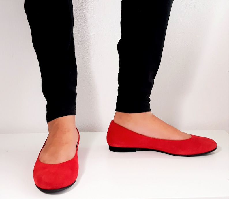 May include: A pair of red suede ballet flats with a pointed toe. The flats are worn by a person wearing black pants.