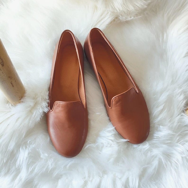 Puede incluir: Un par de mocasines de cuero marr&oacute;n. Los zapatos tienen una punta redondeada y un perfil bajo. Los mocasines se exhiben sobre una superficie blanca y esponjosa. Los zapatos son de color neutro.