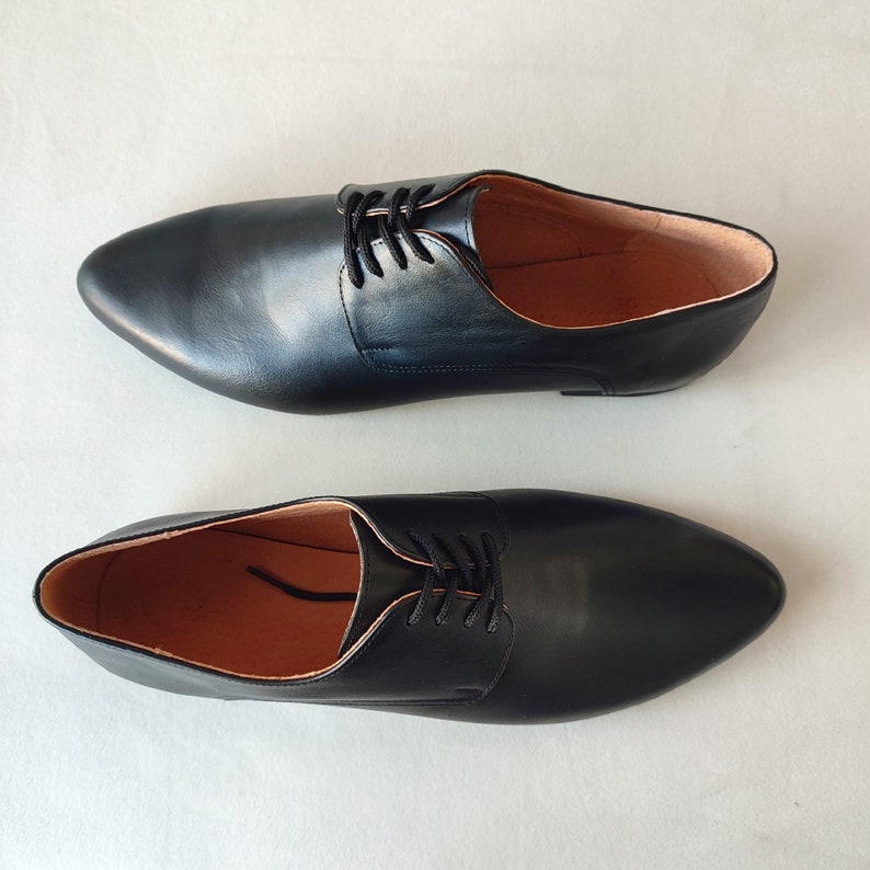Puede incluir: Un par de zapatos negros de cuero con cordones y punta puntiaguda. Los zapatos tienen un dise&ntilde;o sencillo y est&aacute;n hechos de cuero liso.