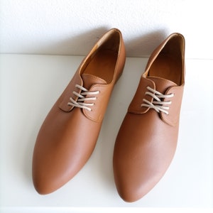 May include: A pair of brown leather oxford shoes with beige laces. The shoes have a classic design with a rounded toe and a low heel. The shoes are displayed on a white surface.