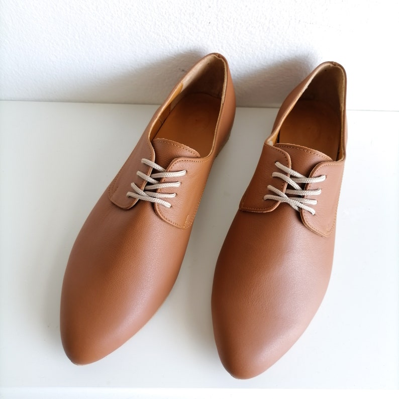 May include: A pair of brown leather oxford shoes with beige laces. The shoes have a classic design with a rounded toe and a low heel. The shoes are displayed on a white surface.