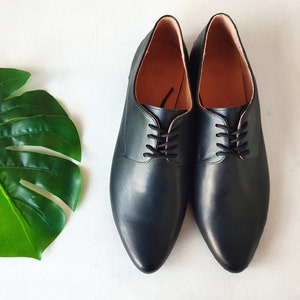 May include: A pair of black leather Oxford shoes with black laces. The shoes have a pointed toe and a smooth, polished finish. The interior is a light brown color. The shoes are displayed on a white surface.