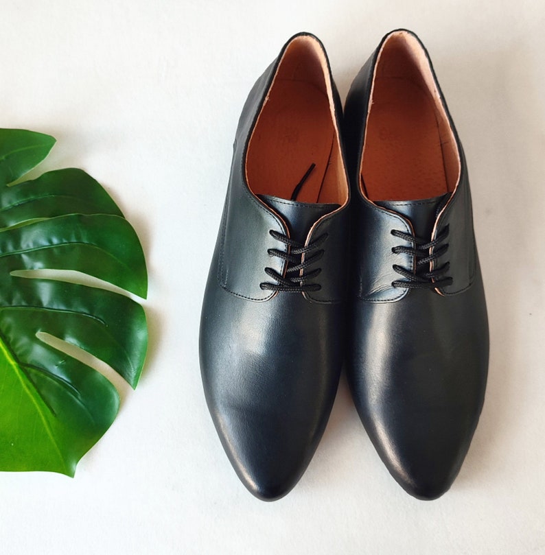 May include: A pair of black leather Oxford shoes with black laces. The shoes have a pointed toe and a smooth, polished finish. The interior is a light brown color. The shoes are displayed on a white surface.