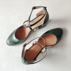 May include: A pair of dark green leather Mary Jane style shoes with ankle straps and small heels. The shoes have a rounded toe and a strap that buckles on the side. The interior of the shoe is a light tan color.
