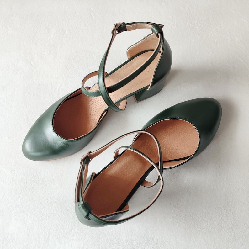 May include: A pair of dark green leather Mary Jane style shoes with ankle straps and small heels. The shoes have a rounded toe and a strap that buckles on the side. The interior of the shoe is a light tan color.