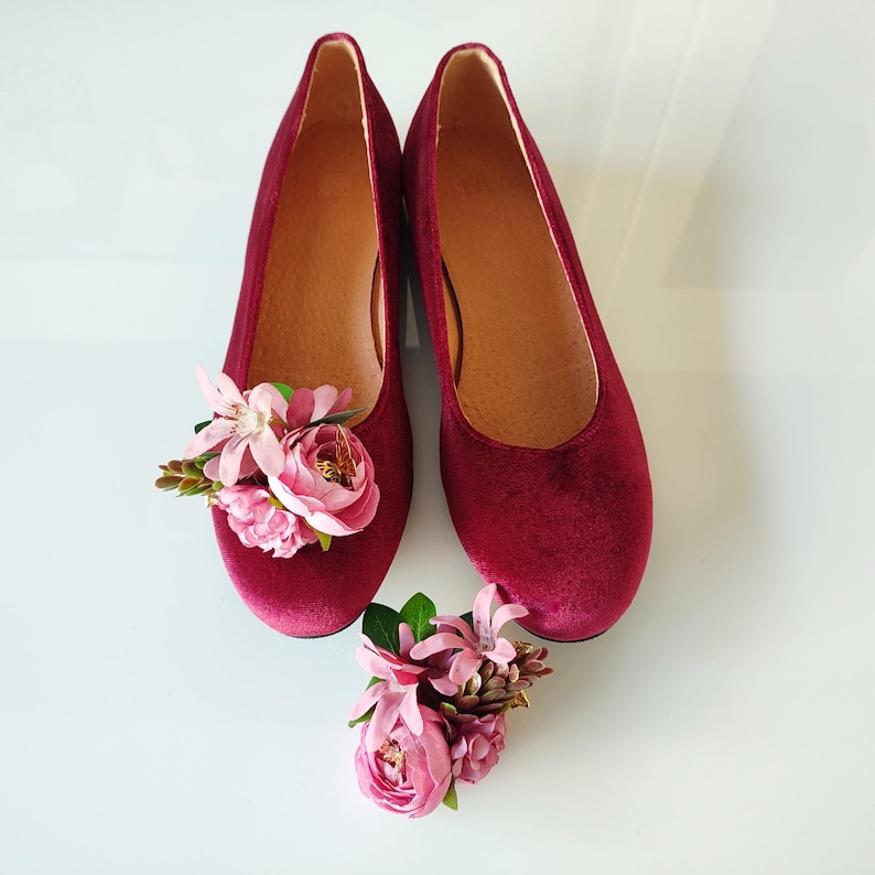 May include: A pair of burgundy velvet flats with pink flower embellishments. The shoes are on a white surface.