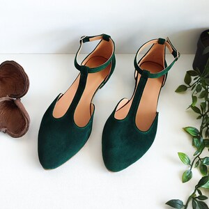May include: A pair of emerald green suede flats with a T-strap design and ankle straps. The shoes have a pointed toe and a small gold buckle. The interior is a light tan color.