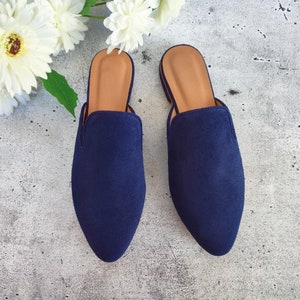 May include: A pair of navy blue suede slip-on mules with a pointed toe and tan insoles. The shoes are displayed on a gray textured surface, with white flowers in the background.