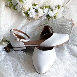 May include: A pair of white leather ankle strap flats with a pointed toe. The shoes are on a white lace fabric background.