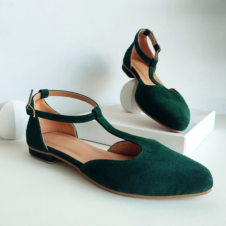 May include: A pair of emerald green suede T-strap flats. The shoes have a rounded toe, a thin ankle strap with a gold buckle, and a low heel. The interior is a light tan color. One shoe is positioned in front of the other.