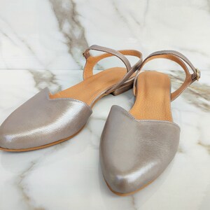 Op de afbeelding: Een paar zilveren leren sandalen met enkelbandjes. De sandalen hebben een spitse neus en een lage hak.