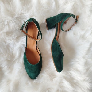 K&ouml;nnte beinhalten: Ein Paar dunkelgr&uuml;ne Wildlederschuhe mit Kn&ouml;chelriemen. Die Schuhe haben eine spitze Zehenpartie und einen niedrigen Blockabsatz.