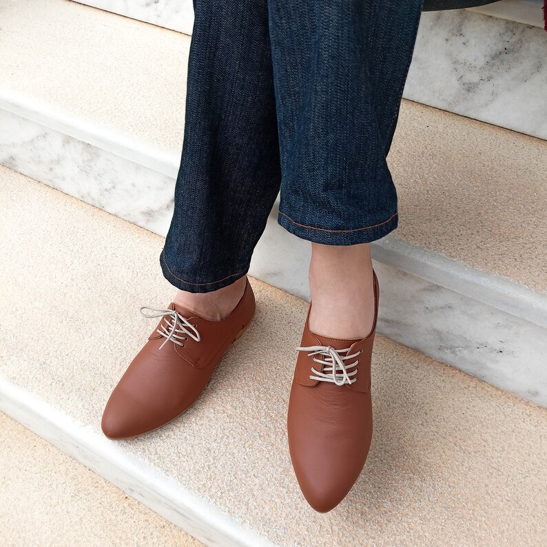 May include: Brown leather oxford shoes with white laces. The shoes have a pointed toe and are worn with blue denim jeans. The shoes are on a white carpeted surface.