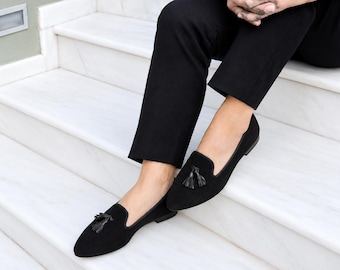 May include: A pair of black suede loafers featuring a pointed toe and a decorative tassel detail on the top of each shoe. The loafers have a low heel and a slim silhouette, paired with black trousers that reach just above the ankle.