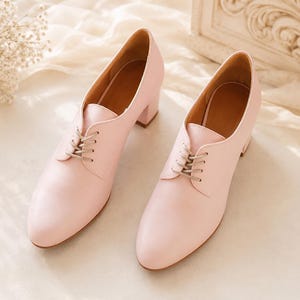 May include: A pair of light pink leather oxford-style shoes with a low block heel. The shoes have a lace-up closure and a pointed toe. The interior of the shoe is a light brown color. The shoes are displayed on a cream-colored fabric.