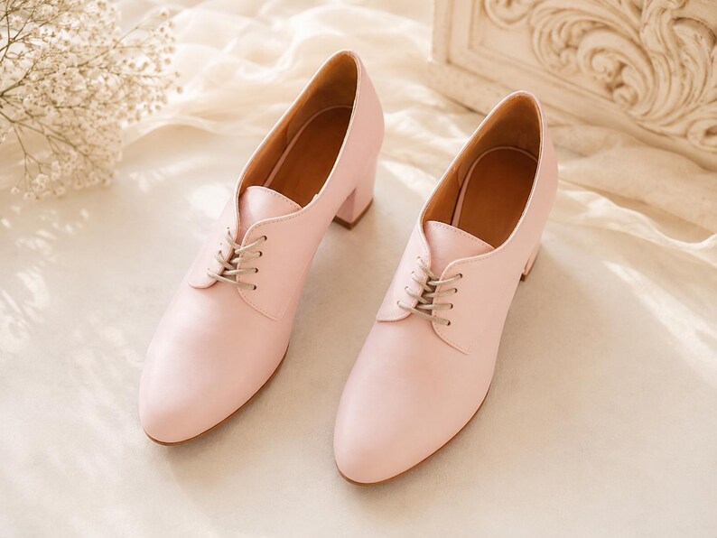 May include: A pair of light pink leather oxford-style shoes with a low block heel. The shoes have a lace-up closure and a pointed toe. The interior of the shoe is a light brown color. The shoes are displayed on a cream-colored fabric.