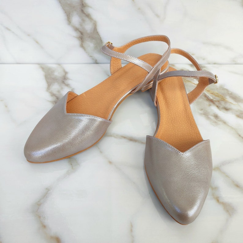 Op de afbeelding: Een paar zilveren metallic leren sandalen met enkelbandjes. De sandalen hebben een spitse neus en een lage hak.