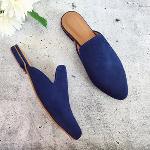May include: A pair of navy blue suede slip-on mules with a pointed toe and a tan insole. The shoes are displayed on a light gray surface with white flowers in the background. The mules are a stylish footwear option.