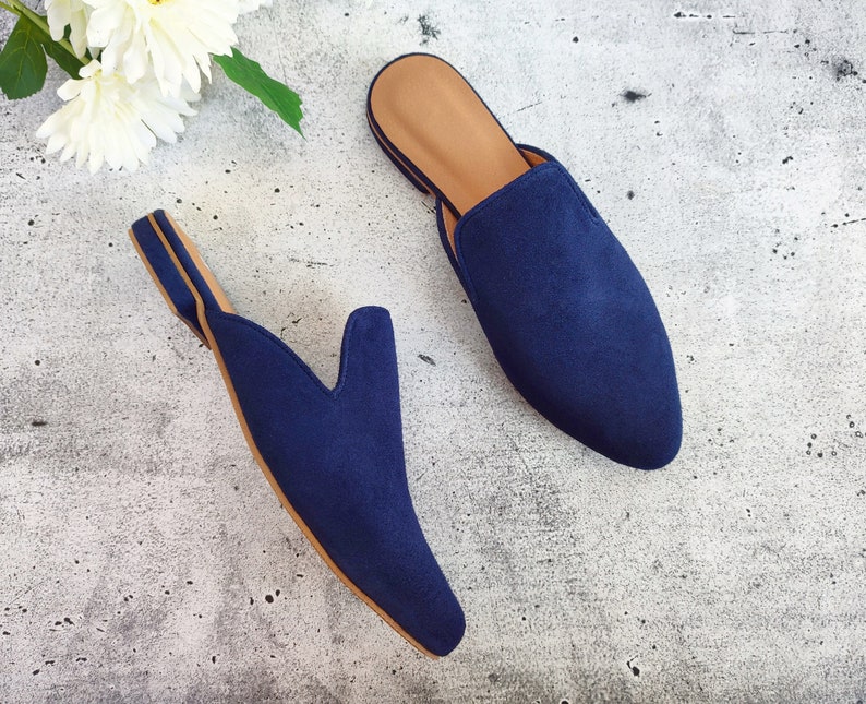 May include: A pair of navy blue suede slip-on mules with a pointed toe and a tan insole. The shoes are displayed on a light gray surface with white flowers in the background. The mules are a stylish footwear option.