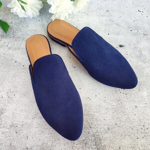 May include: A pair of navy blue suede slip-on mules. The shoes have a pointed toe design and a tan insole. The mules are placed on a light gray surface, with white flowers in the background.