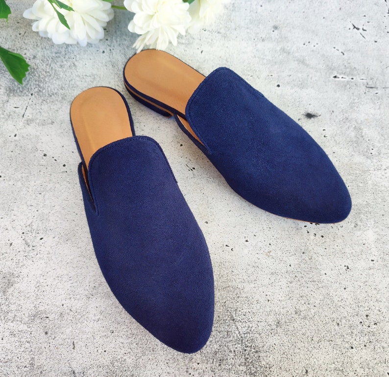 May include: A pair of navy blue suede slip-on mules. The shoes have a pointed toe design and a tan insole. The mules are placed on a light gray surface, with white flowers in the background.