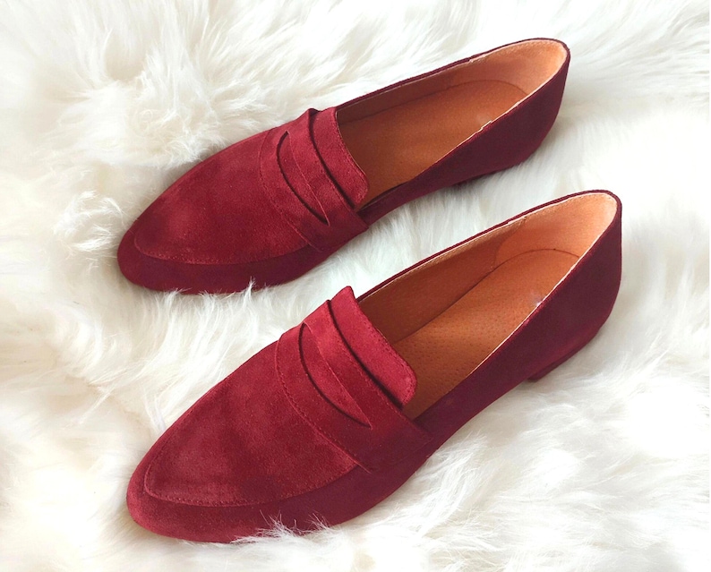May include: A pair of burgundy suede loafers with a pointed toe and a single strap across the vamp.